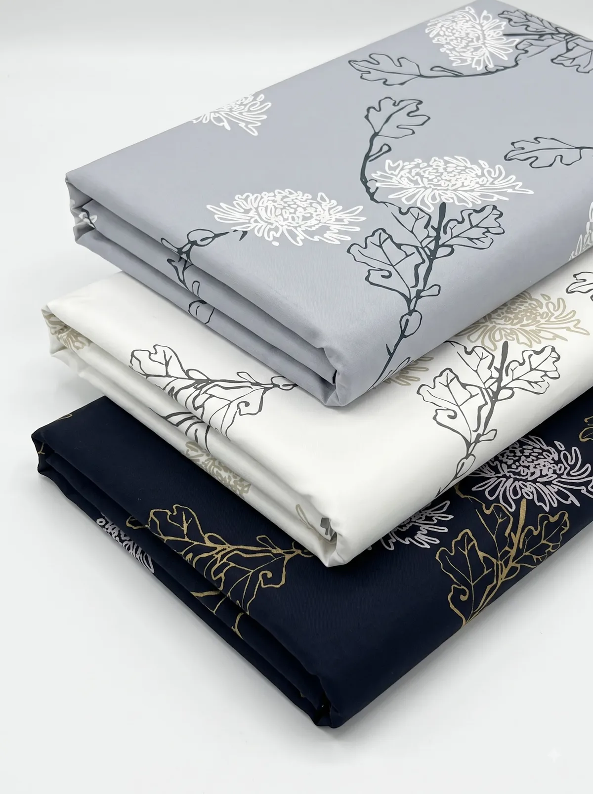 Folded grey, ivory and navy chrysanthemum and oak leaf print fabrics stacked on a white background